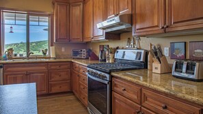 A kitchen with wooden cabinets, a stove, a toaster, and a window with a view of greenery. - The J's Nest Deluxe by Avantstay Sleeps 18 Hot Tub + Game Room Paso Robles (Bradley)