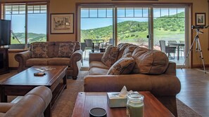 A spacious living room with leather sofas, a coffee table, and a television. Large windows offer a view of a green landscape. - The J's Nest Deluxe by Avantstay Sleeps 18 Hot Tub + Game Room Paso Robles (Bradley)