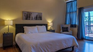 Ein Hotelzimmer mit einem großen Bett, zwei Nachttischen mit Lampen, einem Sessel, einem Bild an der Wand und Blick durch ein mit Vorhängen versehenes Fenster.