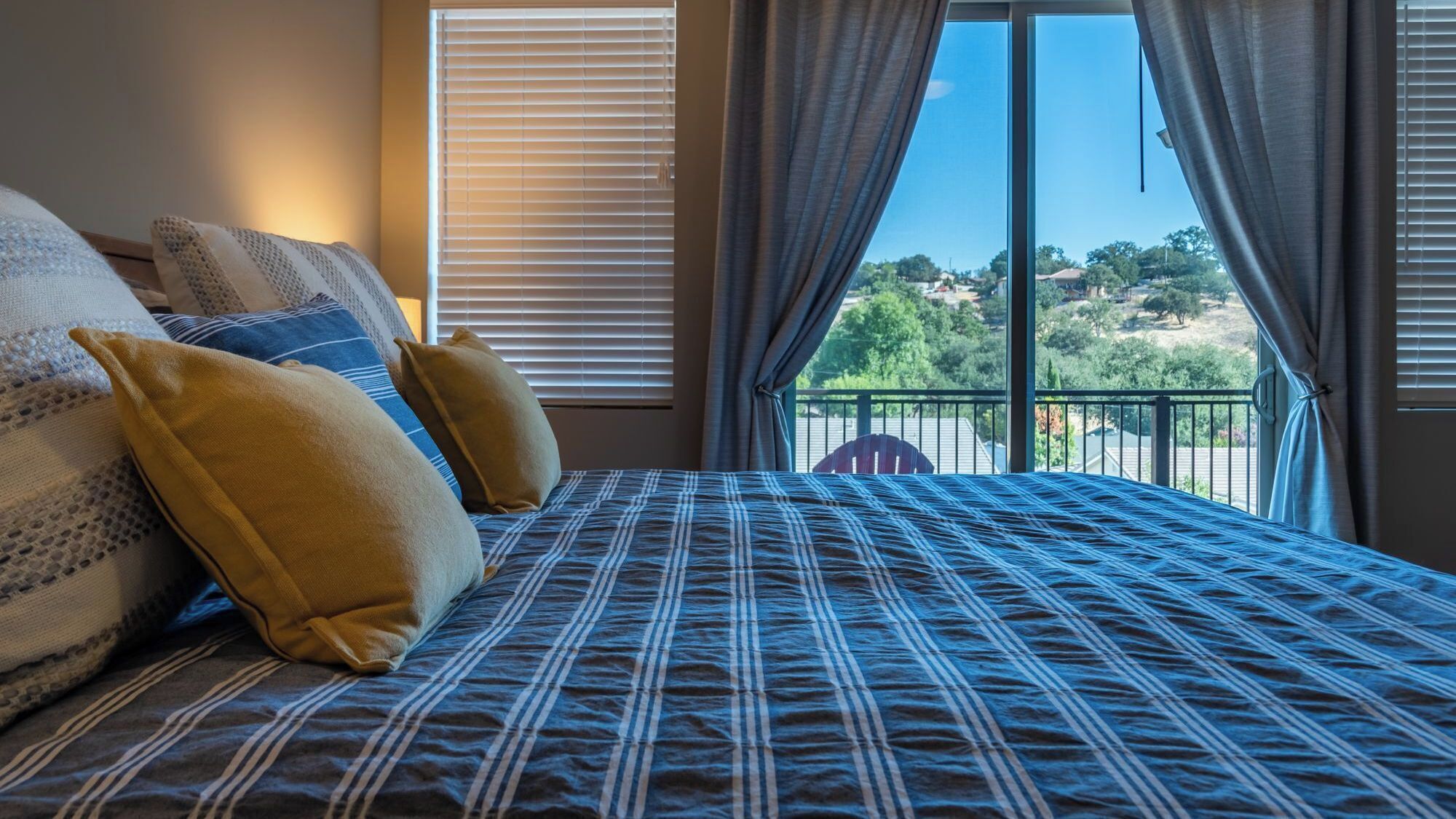 A bedroom with a large bed, two windows with blinds, and a view of a landscape.
