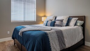 Un dormitorio con cama, almohadas, una colcha, una lámpara de noche y una ventana con persianas.