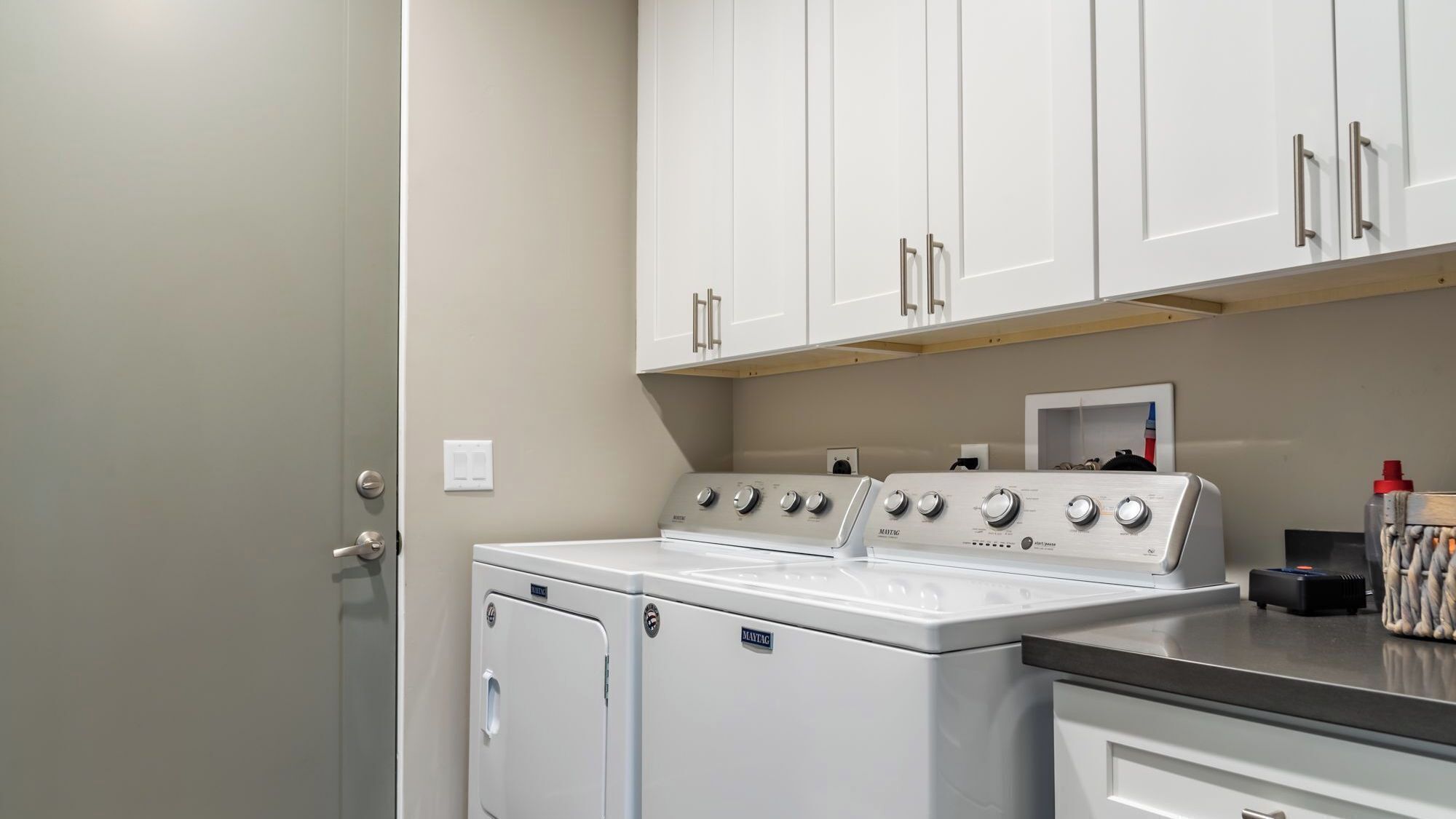 A laundry room with a front-loading washer and dryer, white cabinets above, and a countertop with a kettle and other items.