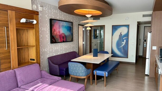 Una habitación de hotel moderna con un sofá púrpura, un armario de madera, una mesa de comedor con sillas y dos grandes cuadros abstractos.
