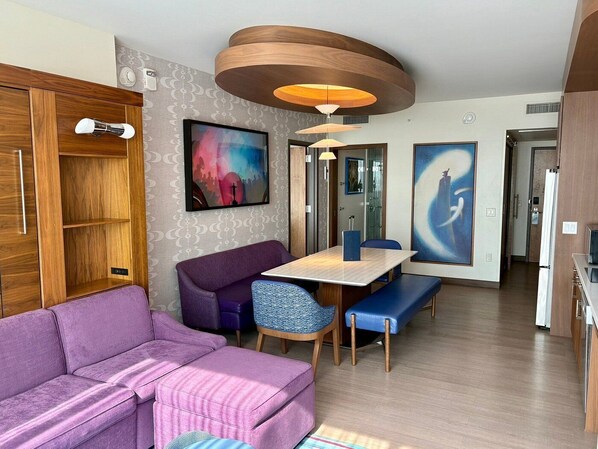A modern hotel room with a purple sofa, a wooden wardrobe, a dining table with chairs, and two large abstract paintings.