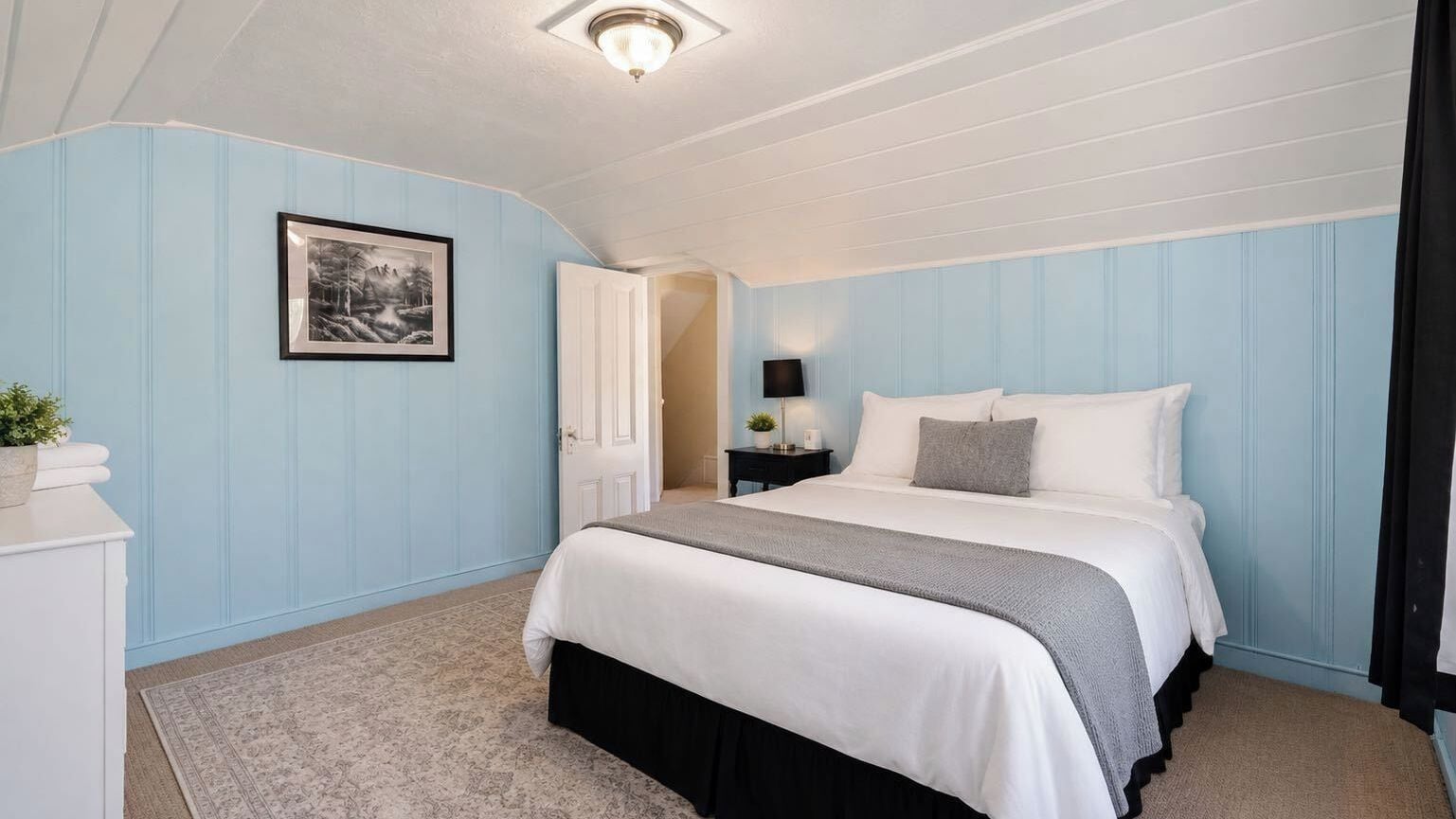 Quarto com cama, mesa de cabeceira, luminária, quadro na parede e porta que dá para outro cômodo.