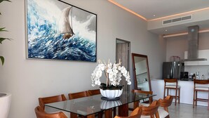 Een moderne eethoek met een glazen tafel en houten stoelen, een groot schilderij met een zeemaand thema, en een keuken op de achtergrond.