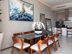 A modern dining area with a glass table and wooden chairs, a large ocean-themed painting, and a kitchen in the background. - Apartment in New Vallarta (Nayarit)