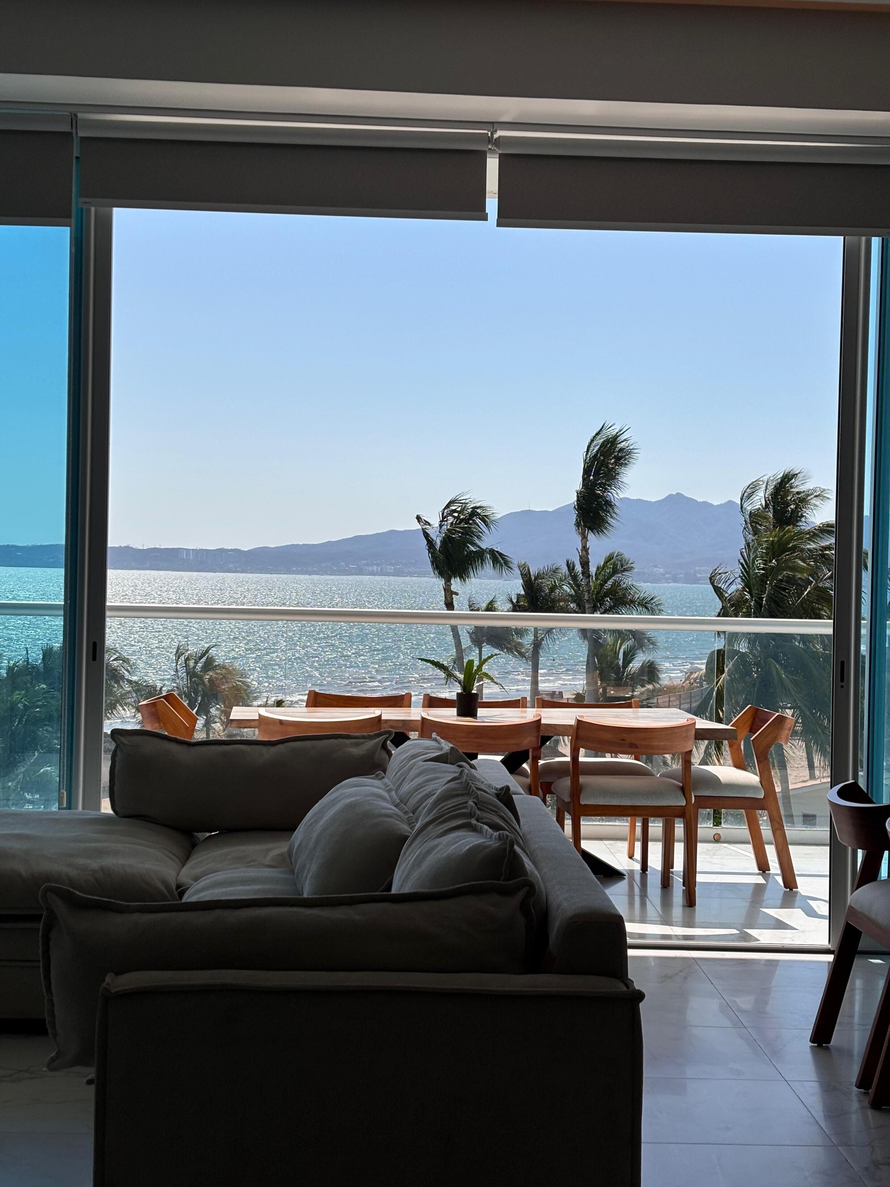 Ein modernes Wohnzimmer mit Sofa und Esstisch, mit Blick auf eine malerische Meereslandschaft mit Palmen und Bergen.