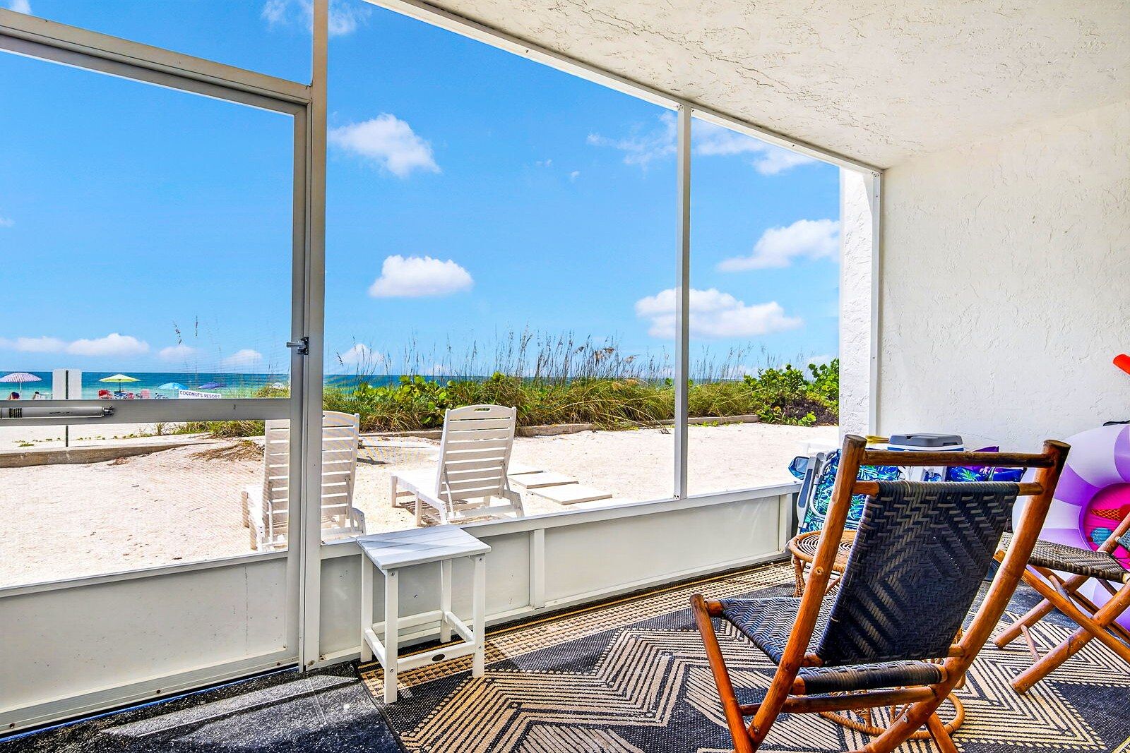 A room with a large window overlooking a beach, a small table, and a wicker chair.