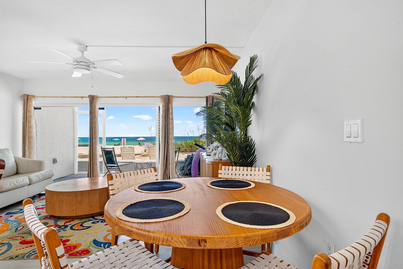 A room with a round wooden table, four chairs, a ceiling fan, and a view of the beach.