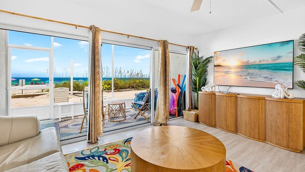A modern living room with a large window overlooking a beach, a round wooden coffee table, and a television on a wooden cabinet.