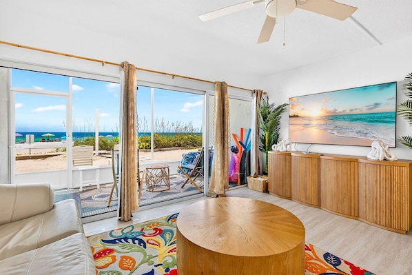 Una sala de estar moderna con un ventanal grande que da a una playa, una mesa redonda de madera y un televisor sobre un mueble de madera.
