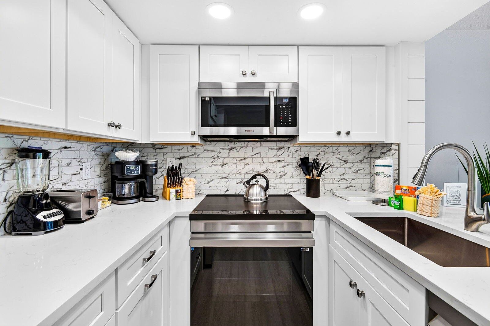 A modern kitchen with white cabinetry, a stainless steel microwave, a kettle, and various appliances.