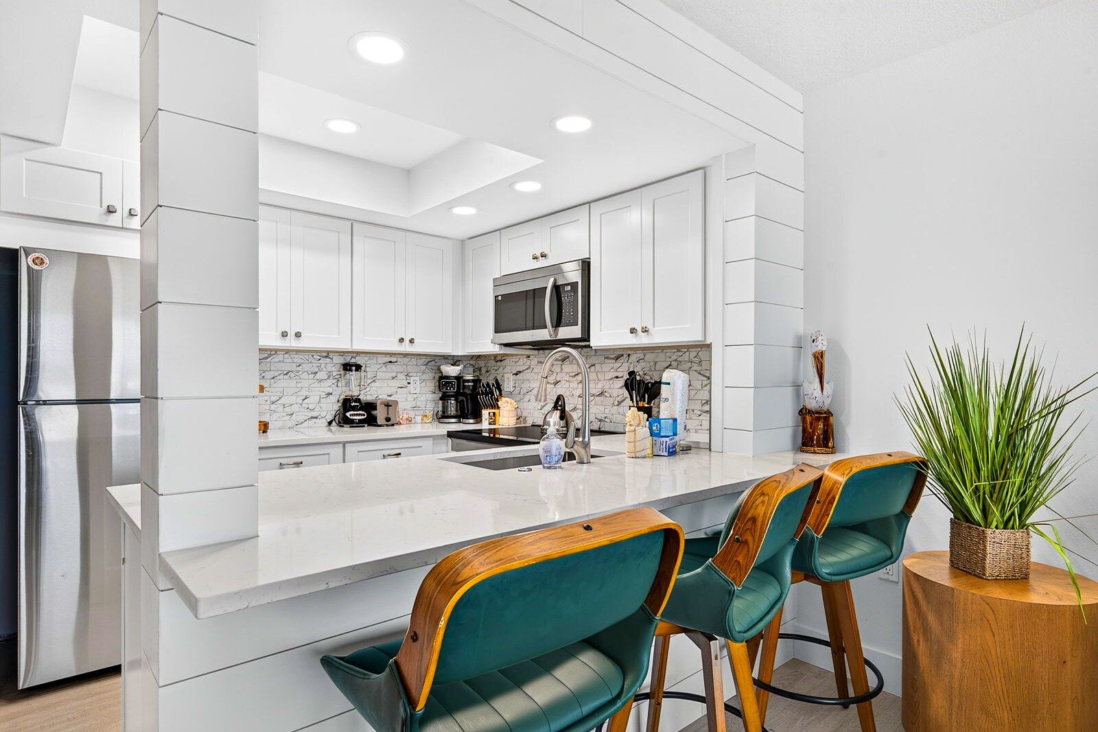 A modern kitchen with white cabinetry, a central island, bar stools, and a potted plant.
