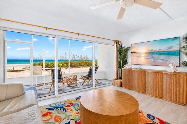 A spacious living room with a large window overlooking a beach, a round wooden coffee table, and a flat-screen TV on the wall.
