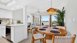 A modern kitchen with white cabinetry, a dining area with a round wooden table and woven chairs, and a view of the beach through large windows.