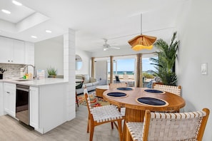Una cocina moderna con armarios blancos, un comedor con una mesa redonda de madera y sillas tejidas, y vista a la playa a través de grandes ventanales.