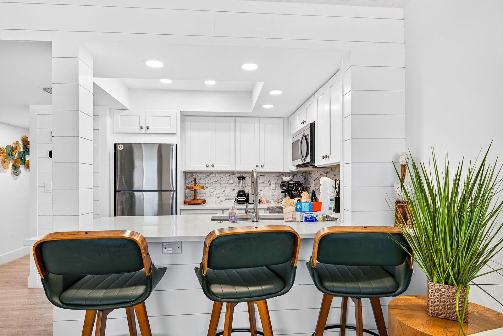 A modern kitchen with white cabinetry, a stainless steel refrigerator, a microwave, and a blender on the counter. There are three bar-style chairs with wooden legs and green upholstery.