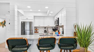 A modern kitchen with white cabinetry, a stainless steel refrigerator, a microwave, and a blender on the counter. There are three bar-style chairs with wooden legs and green upholstery.