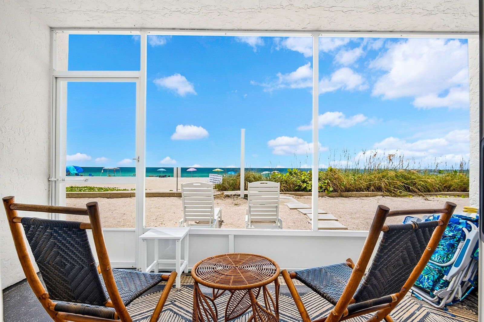 A room with a large window overlooking a beach, two wicker chairs, a small table, and a blue patterned rug.