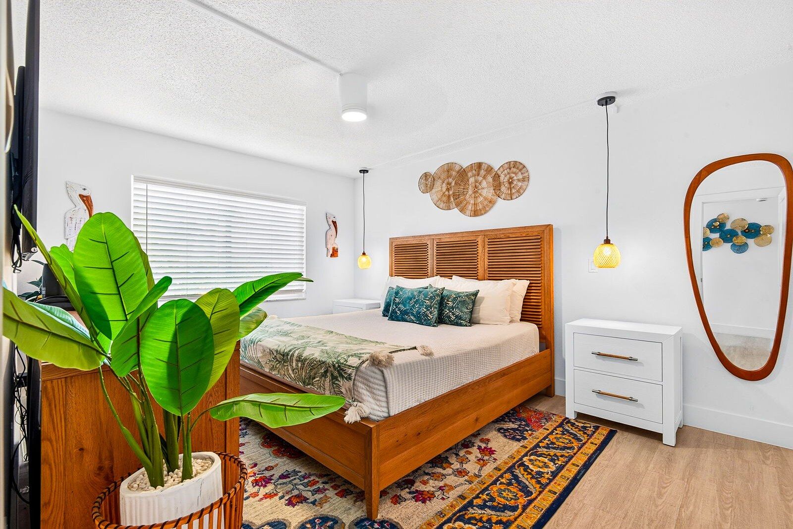A bedroom with a wooden bed, white nightstands, a large potted plant, and a patterned rug.