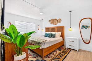 A bedroom with a wooden bed, white nightstands, a large potted plant, and a patterned rug.