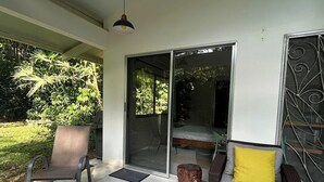 Um pátio externo com uma cadeira, um sofá com almofadas, um banquinho de madeira e uma porta de vidro que dá para um quarto.