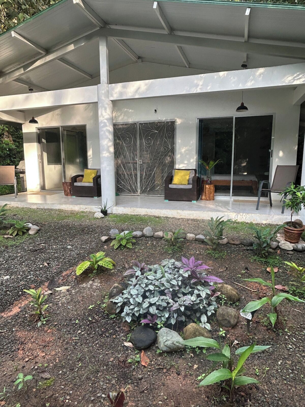 A modern outdoor patio area with a covered seating space, a garden with various plants, and a sliding glass door leading to a covered entrance.