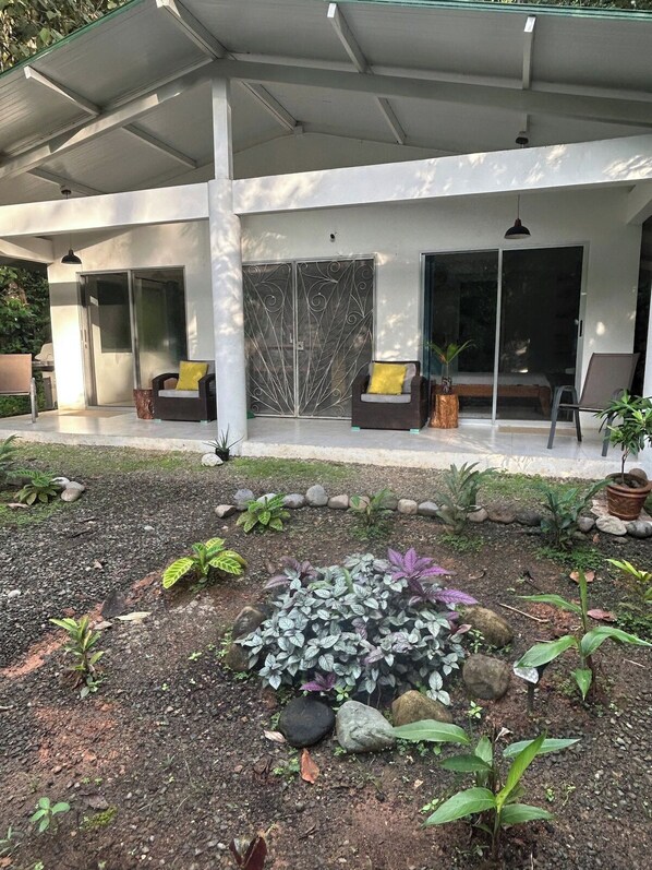 A modern outdoor patio area with a covered seating space, a garden with various plants, and a sliding glass door leading to a covered entrance.