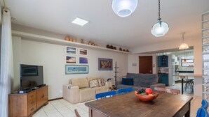 Amplia sala de estar con una mesa de comedor de madera, sillas azules, un sofá y un televisor sobre un mueble de madera.