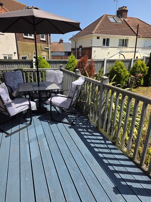 Eine Terrasse mit Tisch und Stühlen, einem schattenspendenden Sonnenschirm und einem Holzgeländer.