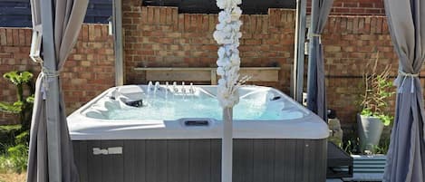 A hot tub under a wooden pergola with curtains, surrounded by potted plants.