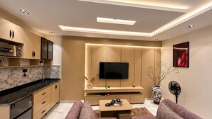 A modern living room with a flat-screen TV, sofa, coffee table, and a kitchen area with cabinets and a stove. - Futuretrendhomes (Abuja)