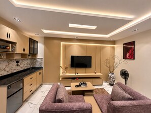 Ein modernes Wohnzimmer mit Flachbildfernseher, Sofa, Couchtisch und einer Küchenzeile mit Schränken und Herd.