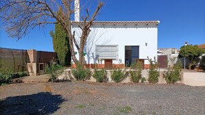 Um edifício branco com telhado de azulejos, chaminé e um pequeno jardim com arbustos e uma árvore.