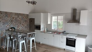 Uma cozinha moderna com armários brancos, uma ilha central com banquetas, micro-ondas, forno e uma janela com persianas.