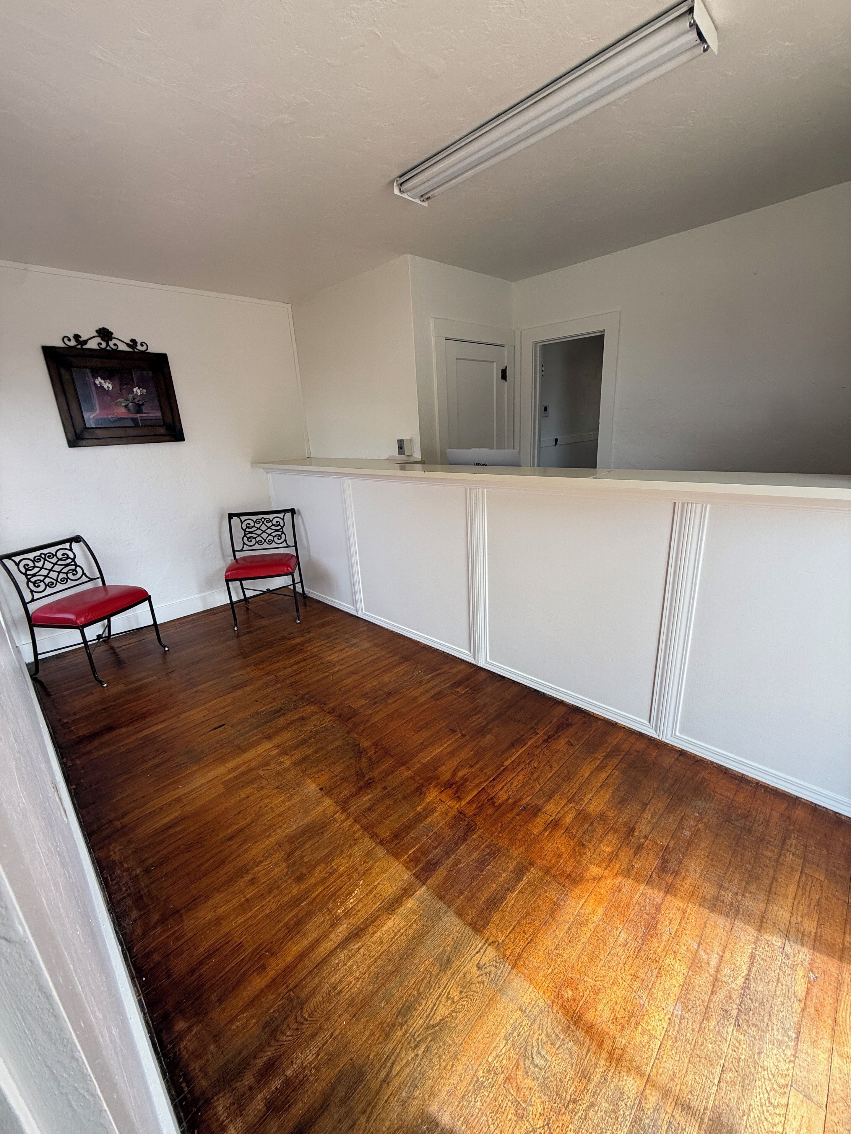 Una habitación con piso de madera, una pared blanca, un cuadro enmarco y dos sillas de metal.