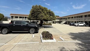 Un estacionamiento con una camioneta negra y un coche blanco, un edificio de techo de tejas, y un árbol en el centro.