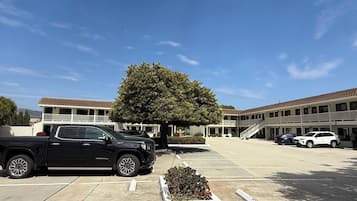 Un estacionamiento con una camioneta negra y un coche blanco, un edificio de techo de tejas, y un árbol en el centro.