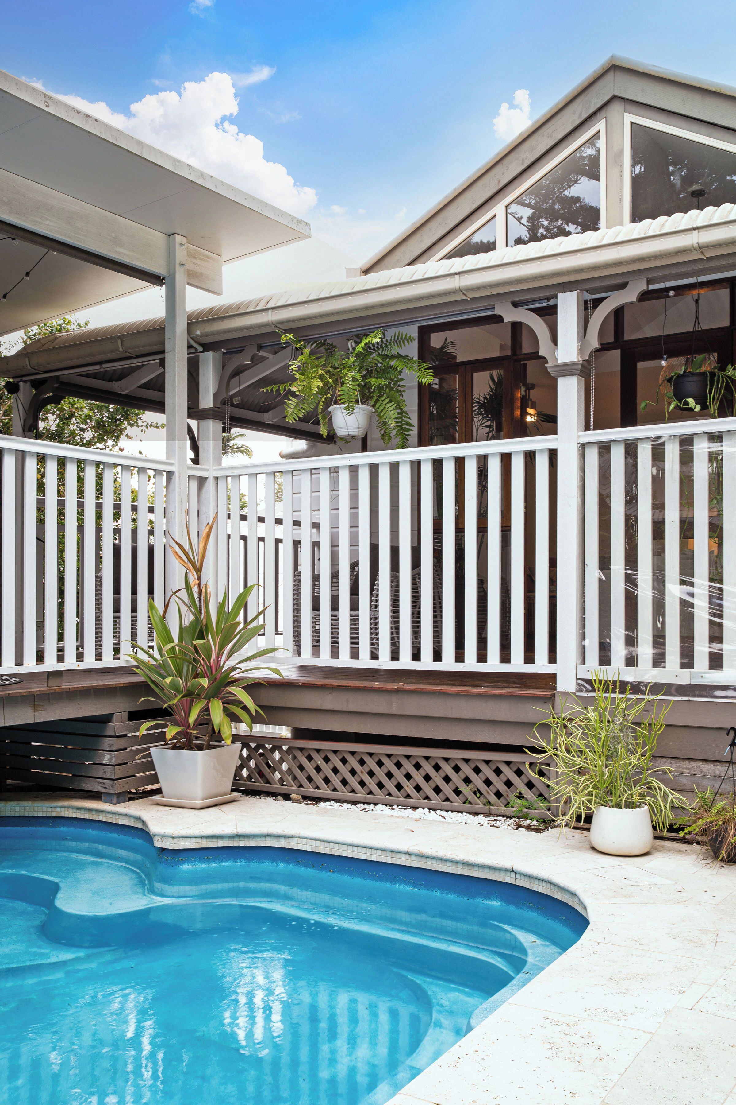 Une piscine dotée d’une terrasse et d’une balustrade blanches, entourée de plantes en pot et d’un patio couvert.