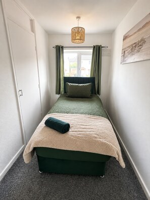 A narrow room with a single bed, a window with curtains, a hanging light fixture, and a painting on the wall. - Spacious 3BR Home | Near Huddersfield Town Centre, University & M62 (Huddersfield)