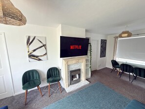 Ein Wohnzimmer mit Kamin, einem Flachbildfernseher, der 'NETFLIX' zeigt, und einer Sitzecke mit zwei grünen Sesseln.