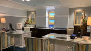 Un espace d’accueil d’hôtel comprenant un comptoir, deux chaises, un bureau avec un ordinateur et un grand miroir.