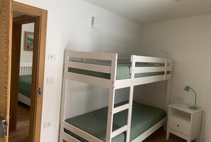 Habitación con literas, una puerta de madera, una mesita de noche con lámpara y un pequeño cajón.