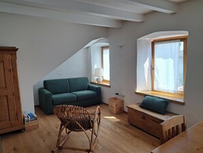 Un espacio tipo loft con un sofá verde, una silla de mimbre, un banco de madera y una ventana con vistas.