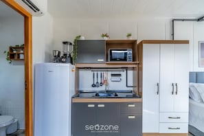 A compact kitchen with a refrigerator, microwave, and coffee maker, a wooden shelf with plants, and a white wardrobe with drawers. - Chalet w/ Parking 200m From the Beach Scj0003 (Florianópolis)