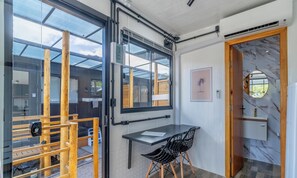 Un interior moderno con una puerta de vidrio que da a un espacio exterior, una pequeña mesa con una silla, y un baño con piso de mármol y un espejo redondo.