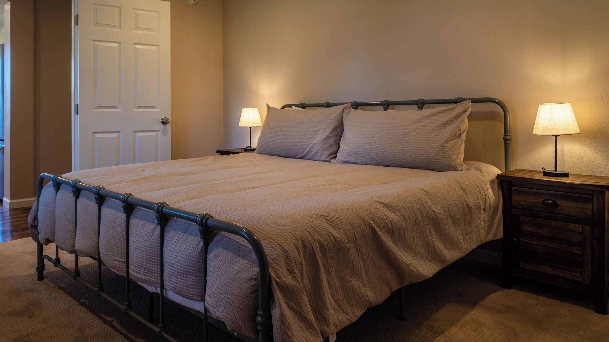 Una cama bien hecha con estructura metálica, dos lámparas de noche y una mesita de noche de madera con cajón.