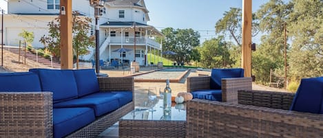 Un'area salotto esterna con mobili in vimini e cuscini blu, un tavolo con piano in vetro, bicchieri da vino e una bottiglia, e vista su una casa con portico e mobili da giardino.