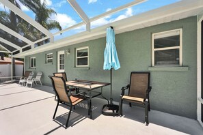 Eine Terrasse im Freien mit Tisch und Stühlen, einem blauen Sonnenschirm und einem überdachten Bereich mit Fenstern.
